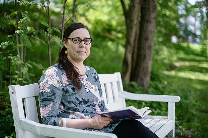 Katja Jokiniemi sitter på en parkbänk med en bok i famnen och tittar in i kameran.