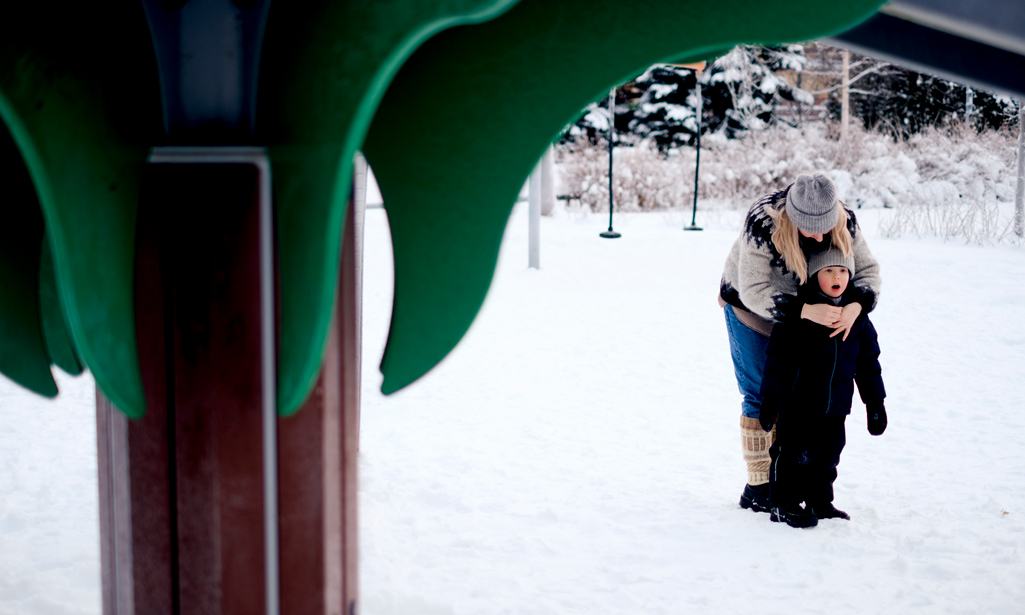 Äiti ja lapsi ovat talvella ulkona leikkipuistossa. Äiti vetää lapsen toppahaalarin vetoketjua kiinni.