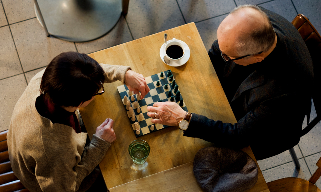 Iäkäs mies ja nainen pelaavat kahvilassa shakkia. Kuva on otettu ylhäältä päin.