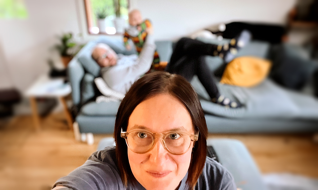 Hanna Hietamäki tar en selfie på sig själv. I bakgrunden ligger maken på soffan och håller babyn i famnen. 