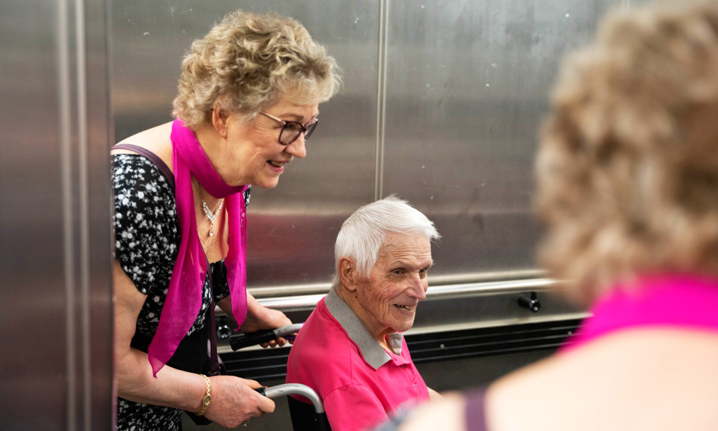 Marja hjälper Arto som sitter i rullstol in i hissen på vårdhemmet. Båda ler glatt.