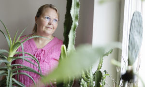 Anne står bakom sina krukväxter och tittar ut genom fönstret.
