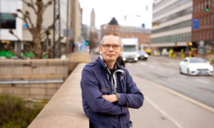 Kari Aro står på gatan och tittar in i kameran. I bakgrunden syns en livligt trafikerad gata.