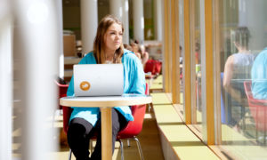 Elena sitter vid ett fönsterbord i biblioteket. Hon har en dator framför sig.