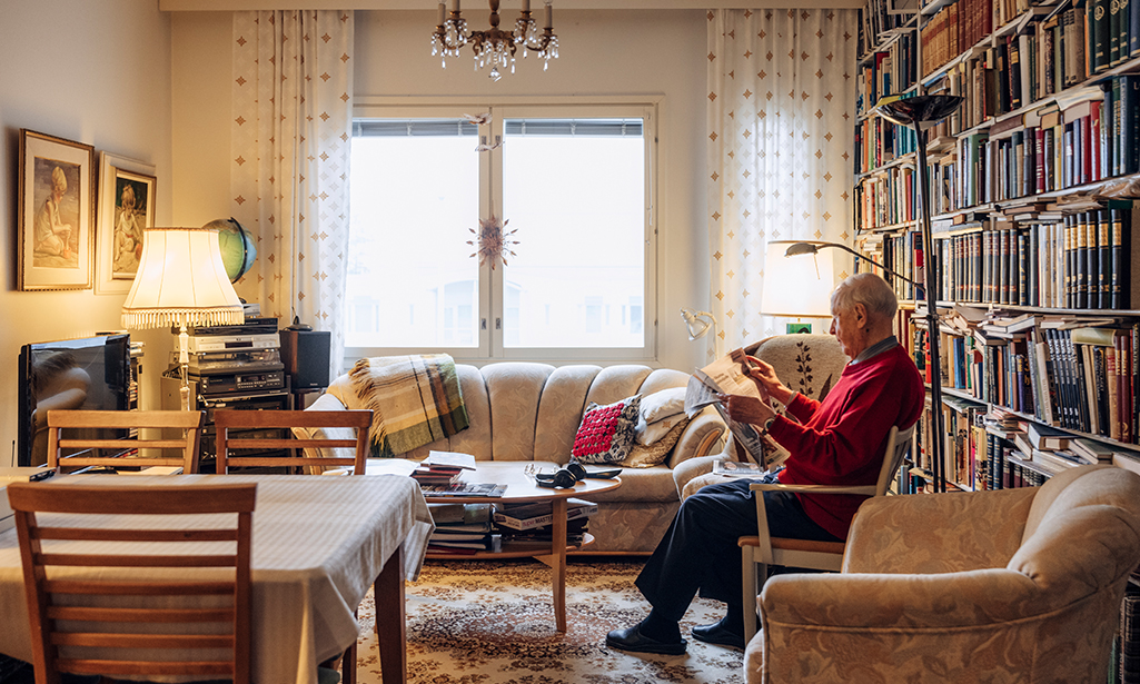 Aimo sitter hemma i en fåtölj och läser en tidning. 