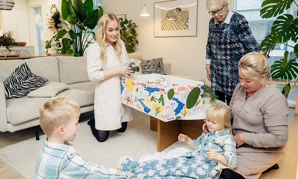 Iida sitter på knä på golvet och undersöker innehållet i moderskapsförpackningen. Hennes mamma och mormor är med. Iidas söner leker också på golvet. 