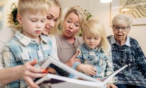 : Iida Latva, Maija Latva och Paula Lehtinen sitter i soffan. Iidas söner sitter i Iidas och Maijas famn. De lyssnar på en saga.