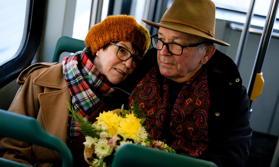 Iäkäs pariskunta istuu raitiovaunussa kukkakimppu kädessään.