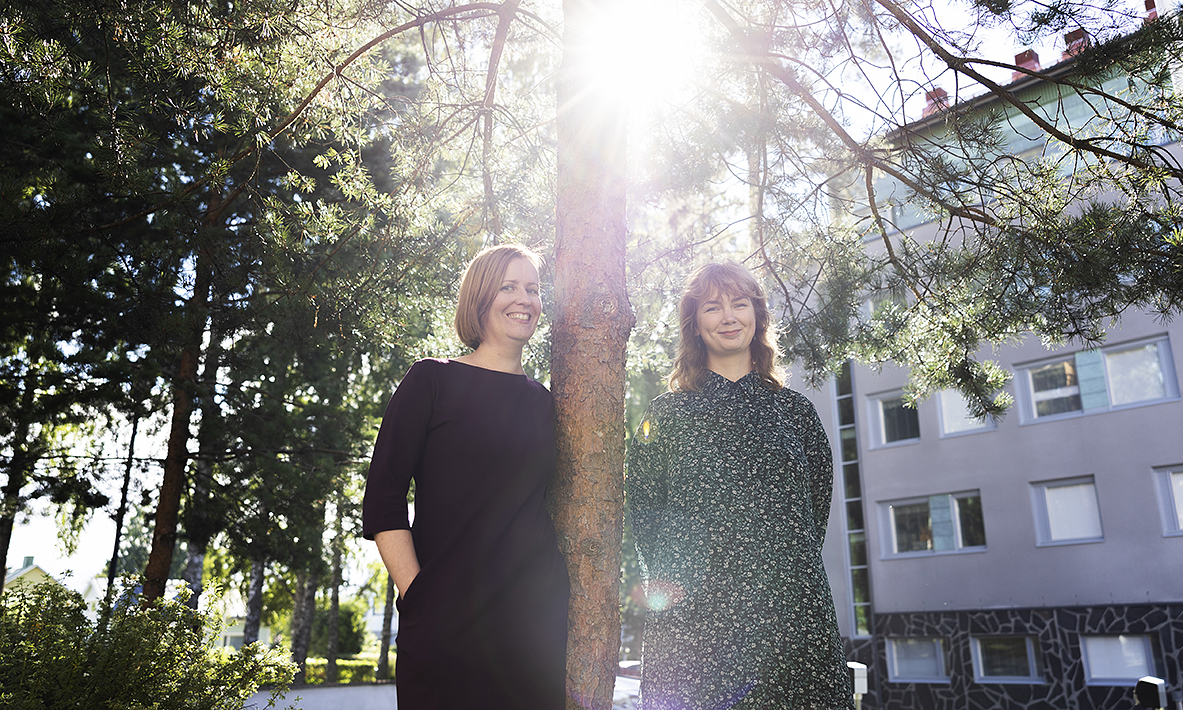 Terveyssosiaalityöntekijät Päivi Rasinmäki (vas.) ja Jenni Hiltunen seisovat ulkona. Aurinko paistaa ja taustalla näkyy sairaalaa.