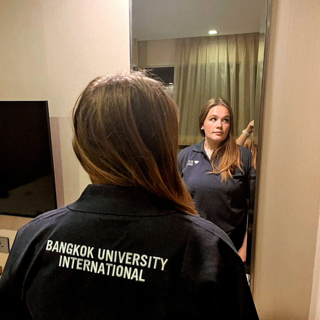 Elena står framför spegeln. Hon är klädd i en skjorta från universitetet i Bangkok med texten Bangkok University International på ryggen.